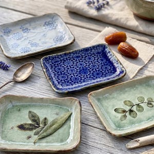 May include: Four small, rectangular ceramic dishes with decorative patterns and designs. One dish has a blue floral pattern, another has a blue geometric pattern, and two have green and brown leaf designs. A silver spoon and lavender sprigs are also present.