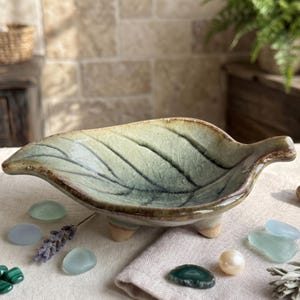 May include: A ceramic leaf-shaped dish with a green and brown glaze, resting on small feet. The dish is surrounded by decorative stones, a sprig of lavender, and a pearl. The dish is approximately 8 inches long.
