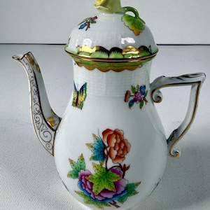 May include: A white porcelain teapot featuring a basket-weave texture on the upper body and lid. The teapot is decorated with hand-painted colorful floral motifs, a yellow butterfly, and gold trim. A sculpted yellow rose serves as the lid handle.