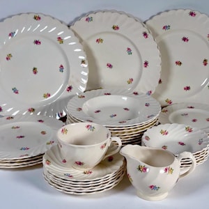 Puede incluir: Un juego de vajilla completo de color crema con un estampado floral. El juego incluye platos, cuencos, tazas, platillos y una jarra de crema. Los platos tienen un borde festoneado y un diseño en espiral. Cada pieza está decorada con pequeños racimos florales coloridos.