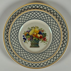 May include: A set of three white ceramic plates with a fruit basket design. The plates have a green lattice pattern around the edges and a yellow trim. The center plate features a basket of colorful fruits.