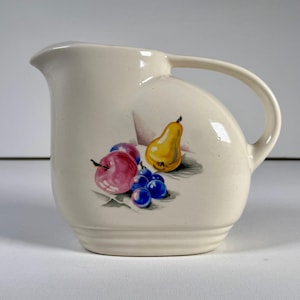 May include: Cream-colored ceramic pitcher with a curved handle and a spout. The pitcher is decorated with a colorful illustration of a yellow pear, red apple, purple plums, and blue grapes. The pitcher has a ribbed base.