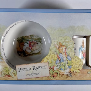 May include: A Peter Rabbit by Wedgwood boxed set featuring a bowl and a mug. The bowl has an illustration of a rabbit and text around the rim. The mug and box feature illustrations of Peter Rabbit. The box has the Peter Rabbit logo.