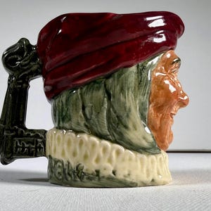 May include: A ceramic mug shaped like a man's head, featuring a burgundy hat, green hair, and an orange face. The mug has a black handle and a cream-colored ruffled collar. The mug is a decorative item.