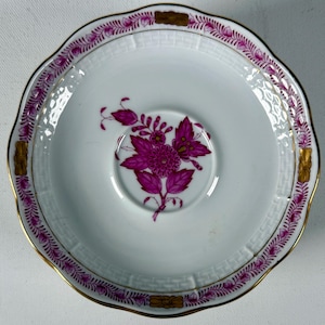 May include: A white porcelain saucer featuring a central magenta floral motif. The rim is decorated with a magenta leaf pattern, gold accents, and a textured basket-weave border. The edge of the saucer has a scalloped shape with a gold trim.