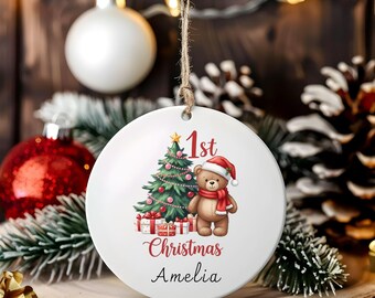 May include: A white Christmas ornament with a festive design. It features a Christmas tree, a teddy bear wearing a Santa hat and scarf, and the text "1st Christmas Amelia" in red and black. The ornament hangs from a twine string.