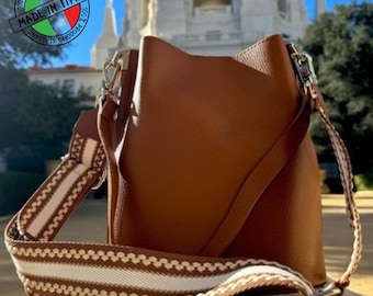 May include: A brown leather bucket bag with a patterned shoulder strap. The bag has a silver-tone clasp and a tag. The strap is white with a brown zigzag pattern. The bag is made in Italy.