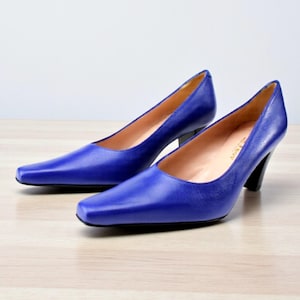 May include: A pair of cobalt blue leather pumps with a square toe and a black heel. The shoes are set on a light wood surface.