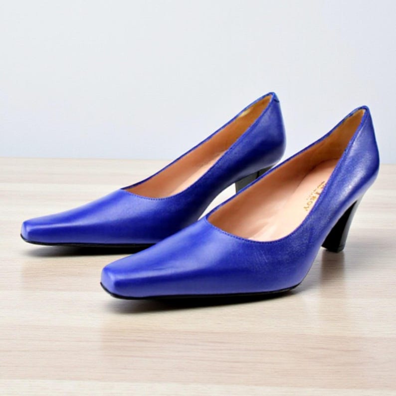 May include: A pair of cobalt blue leather pumps with a square toe and a black heel. The shoes are set on a light wood surface.