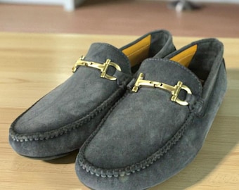 May include: A pair of gray suede loafers with gold-colored metal accents. The shoes have a classic slip-on design with decorative stitching around the edges. The interior lining is yellow.