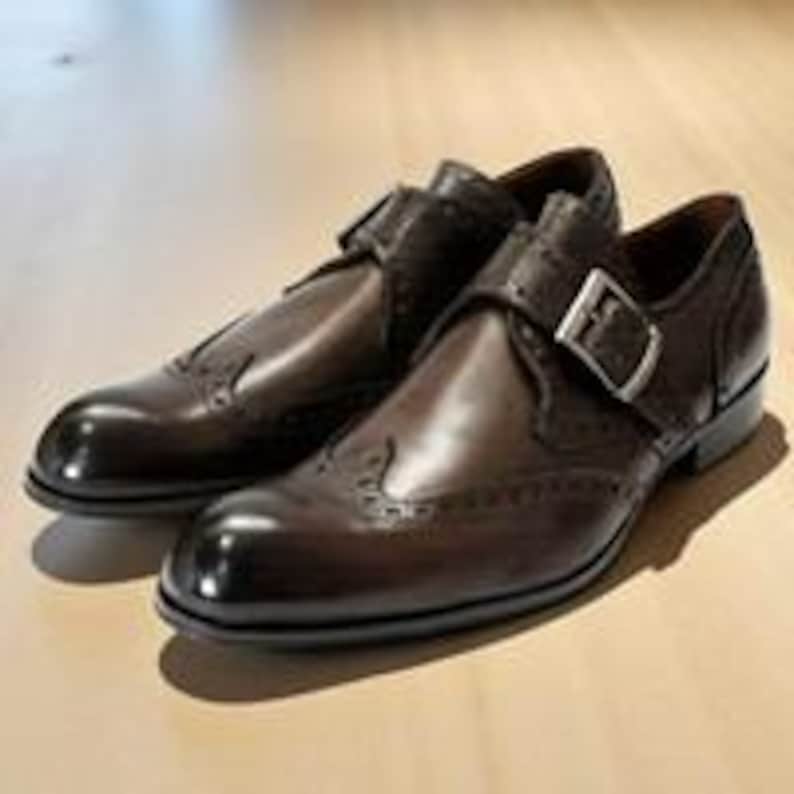 May include: A pair of dark brown leather monk strap shoes featuring a wingtip design with decorative brogue perforations along the seams. Each shoe has a single silver-toned metal buckle closure on the side and a polished finish.