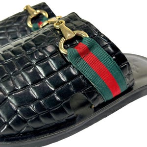 May include: A pair of black slip-on sandals featuring a glossy crocodile-embossed texture. Each sandal is adorned with a gold-toned metal horsebit detail centered over a green and red striped fabric band across the top of the foot.