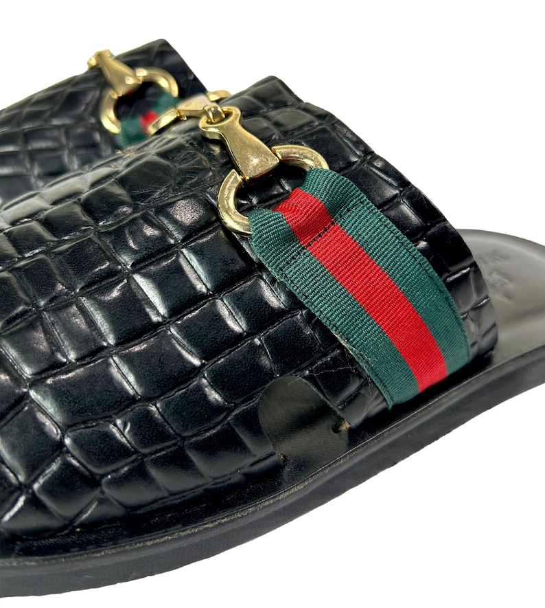 May include: A pair of black slip-on sandals featuring a glossy crocodile-embossed texture. Each sandal is adorned with a gold-toned metal horsebit detail centered over a green and red striped fabric band across the top of the foot.