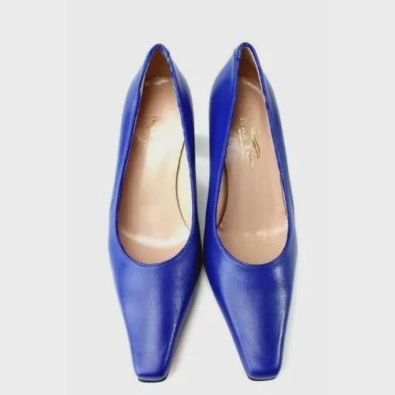 May include: A pair of cobalt blue pumps with a pointed toe and a low heel. The shoes have a smooth, shiny finish and a neutral-colored interior. The shoes are set against a white background.