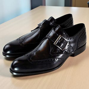 May include: A pair of dark brown leather monk strap shoes featuring a wingtip design with decorative brogue perforations along the seams and a single silver-toned metal buckle closure on the side of each shoe with a polished finish.