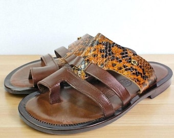 Può includere: Un paio di sandali in pelle marrone con design infradito e una fascia larga sul collo del piede. La fascia superiore è arricchita da un materiale testurizzato con motivo a pelle di serpente arancione e nero e un piccolo emblema metallico dorato.