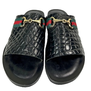 May include: A pair of black slide sandals featuring a textured crocodile-embossed pattern on the upper strap. Each sandal is adorned with a gold-tone metal horsebit detail centered over a horizontal green and red striped fabric band.