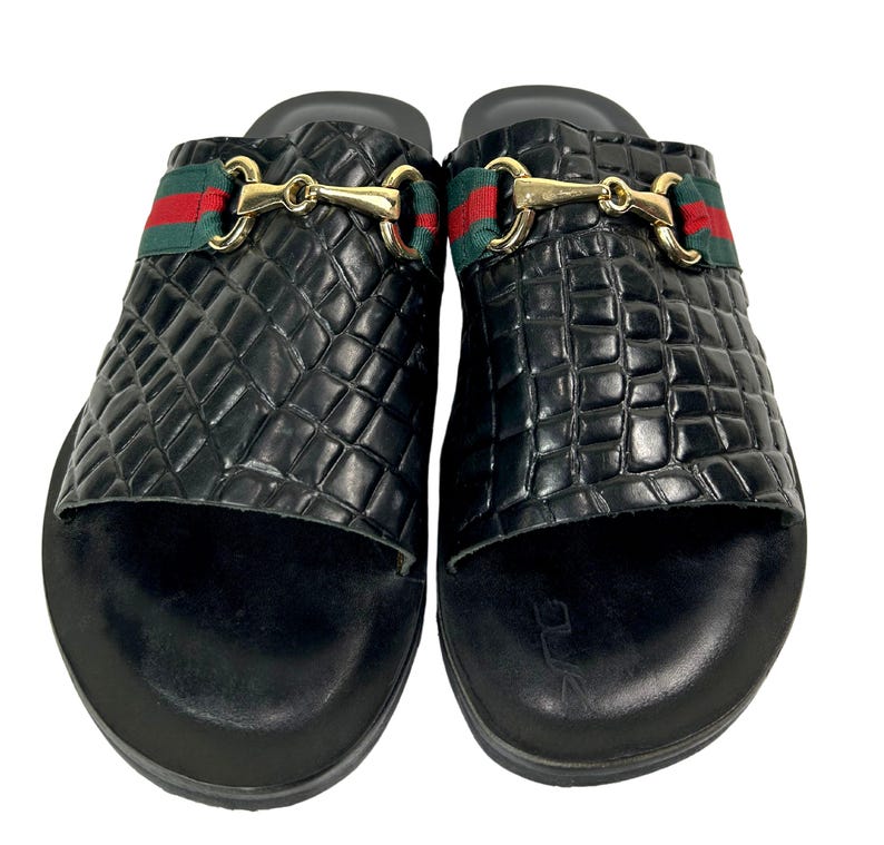 May include: A pair of black slide sandals featuring a textured crocodile-embossed pattern on the upper strap. Each sandal is adorned with a gold-tone metal horsebit detail centered over a horizontal green and red striped fabric band.
