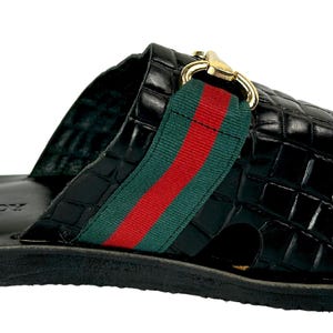 May include: A black slide sandal with a crocodile-embossed texture, featuring a wide strap across the top adorned with a green and red fabric stripe and a gold-toned metal horsebit detail. The footbed is black with a logo embossed on the heel.