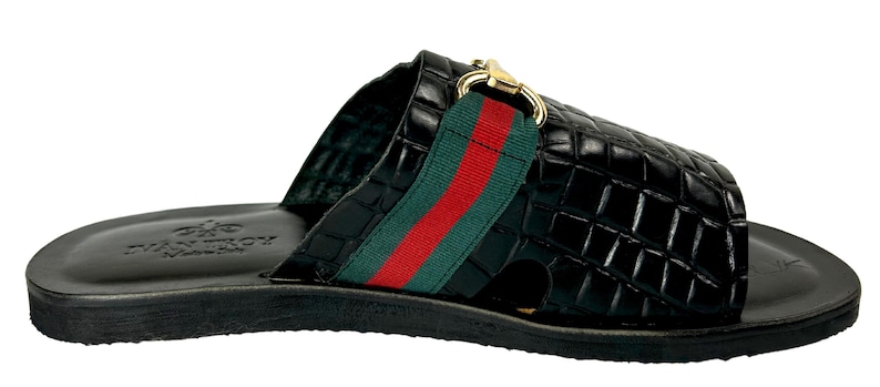 May include: A black slide sandal with a crocodile-embossed texture, featuring a wide strap across the top adorned with a green and red fabric stripe and a gold-toned metal horsebit detail. The footbed is black with a logo embossed on the heel.
