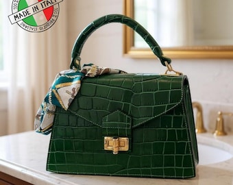 May include: A dark green handbag with a crocodile skin texture, a top handle, and a gold-colored clasp. A patterned scarf is tied to the handle. The bag has a rectangular shape and a flap closure. The text "MADE IN ITALY" is visible.