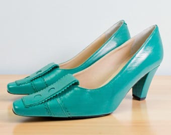 May include: A pair of teal pumps featuring a square toe, a block heel, and a decorative rectangular tab on the vamp with scalloped edges and two circular cutouts. The shoes have a smooth finish and a light-colored interior lining.