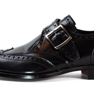 May include: A black leather monk strap shoe featuring a wingtip design with decorative brogue perforations along the seams. The shoe has a polished finish, a silver-toned metal buckle closure, and a stacked heel with a dark sole.