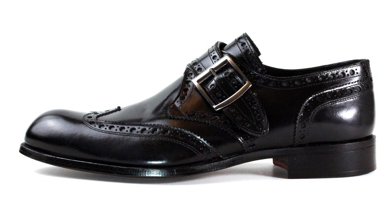 May include: A black leather monk strap shoe featuring a wingtip design with decorative brogue perforations along the seams. The shoe has a polished finish, a silver-toned metal buckle closure, and a stacked heel with a dark sole.