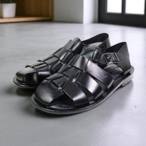 Könnte beinhalten: Ein Paar schwarze Ledersandalen mit geflochtenem Design. Die Sandalen haben eine geschlossene Zehenpartie und einen Schnallenverschluss am Knöchelriemen. Die Schuhe stehen auf einer grauen Oberfläche.