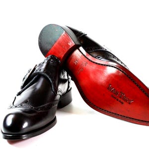 May include: A pair of black leather monk strap shoes with brogue detailing. One shoe is upright, while the other is positioned to show the bright red sole, which features the embossed text 'Mani Troy' and 'Made in Italy' along with the size 42.