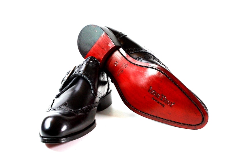 May include: A pair of black leather monk strap shoes with brogue detailing. One shoe is upright, while the other is positioned to show the bright red sole, which features the embossed text 'Mani Troy' and 'Made in Italy' along with the size 42.