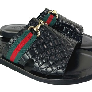 May include: A pair of black slide sandals featuring a textured crocodile-embossed finish, a green and red striped fabric band across the top, and a gold-toned metal horsebit detail. The footbed is smooth black leather with a debossed logo stamp.