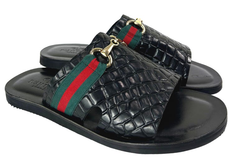 May include: A pair of black slide sandals featuring a textured crocodile-embossed finish, a green and red striped fabric band across the top, and a gold-toned metal horsebit detail. The footbed is smooth black leather with a debossed logo stamp.