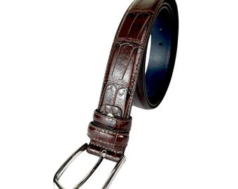 Può includere: Una cintura in pelle marrone con una trama a pelle di coccodrillo. La cintura ha una fibbia color argento e un lato inferiore blu scuro. La cintura è angolata, mostrando il suo design e la sua trama su sfondo bianco.