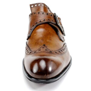 May include: A tan leather monk strap shoe featuring a burnished toe, intricate brogue detailing along the seams, and a single metallic buckle closure. The shoe has a polished finish and a dark sole, showcasing a classic design with visible stitching.