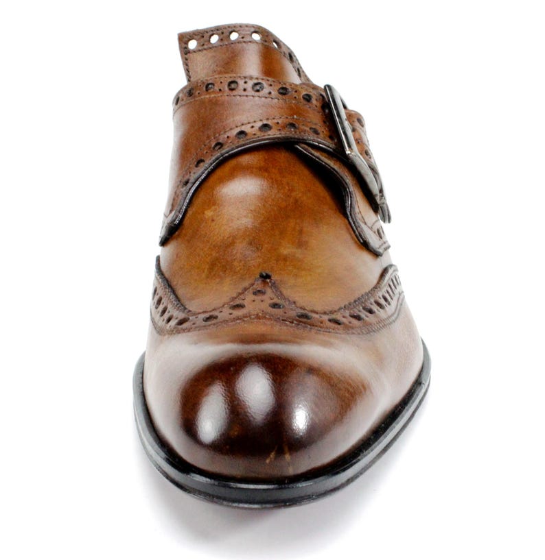 May include: A tan leather monk strap shoe featuring a burnished toe, intricate brogue detailing along the seams, and a single metallic buckle closure. The shoe has a polished finish and a dark sole, showcasing a classic design with visible stitching.