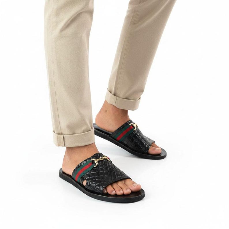 May include: Black leather slide sandals featuring a textured crocodile-embossed finish, a horizontal green and red fabric stripe across the strap, and a gold-toned metal horsebit hardware detail. The sandals have a flat black sole and open toe.