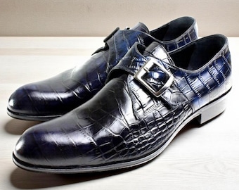 May include: A pair of navy blue leather dress shoes with a crocodile skin texture. Each shoe features a silver buckle closure and a low heel. The shoes are displayed on a light wooden surface.