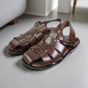 Könnte beinhalten: Ein Paar braune Ledersandalen mit geflochtenem Design. Die Sandalen haben eine geschlossene Zehenpartie und einen Knöchelriemen mit Schnalle. Das Leder ist glatt und glänzend. Ein stilvoller Sommerschuh.