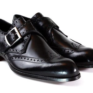 May include: A pair of black leather monk strap shoes featuring a polished finish, wingtip brogue detailing, and a silver-toned metal buckle closure on the side strap. The shoes have a rounded toe and a low heel, showcasing a classic design.