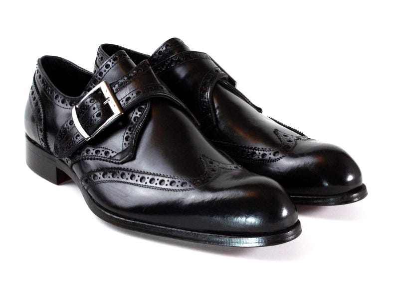 May include: A pair of black leather monk strap shoes featuring a polished finish, wingtip brogue detailing, and a silver-toned metal buckle closure on the side strap. The shoes have a rounded toe and a low heel, showcasing a classic design.