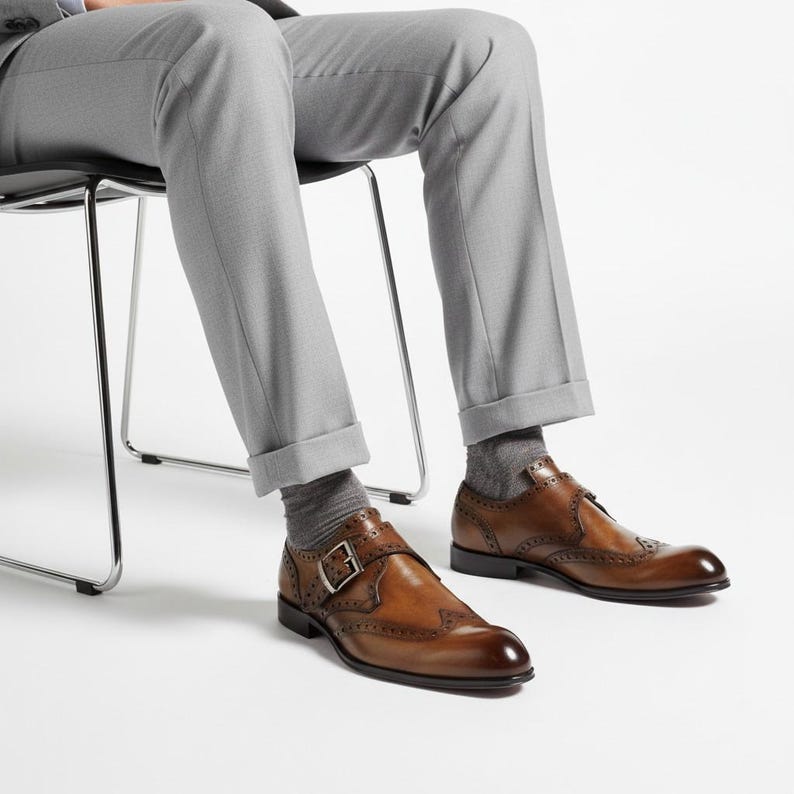 May include: A pair of brown leather monk strap shoes featuring brogue detailing, a silver-toned metal buckle, and a rounded toe. The shoes are worn with grey socks and light grey trousers, showcasing a classic design with decorative perforations.