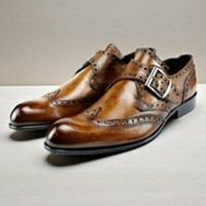 May include: A pair of brown leather monk strap shoes featuring a burnished finish, intricate brogue detailing along the seams, a pointed toe, and a silver-toned metal buckle closure on the side strap, set on a dark sole with a low heel.