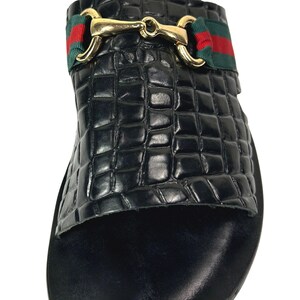 May include: A black slide sandal featuring a textured crocodile-embossed finish on the upper strap. The strap is accented with a horizontal green and red fabric band and a polished gold-tone metal horsebit hardware detail across the center.