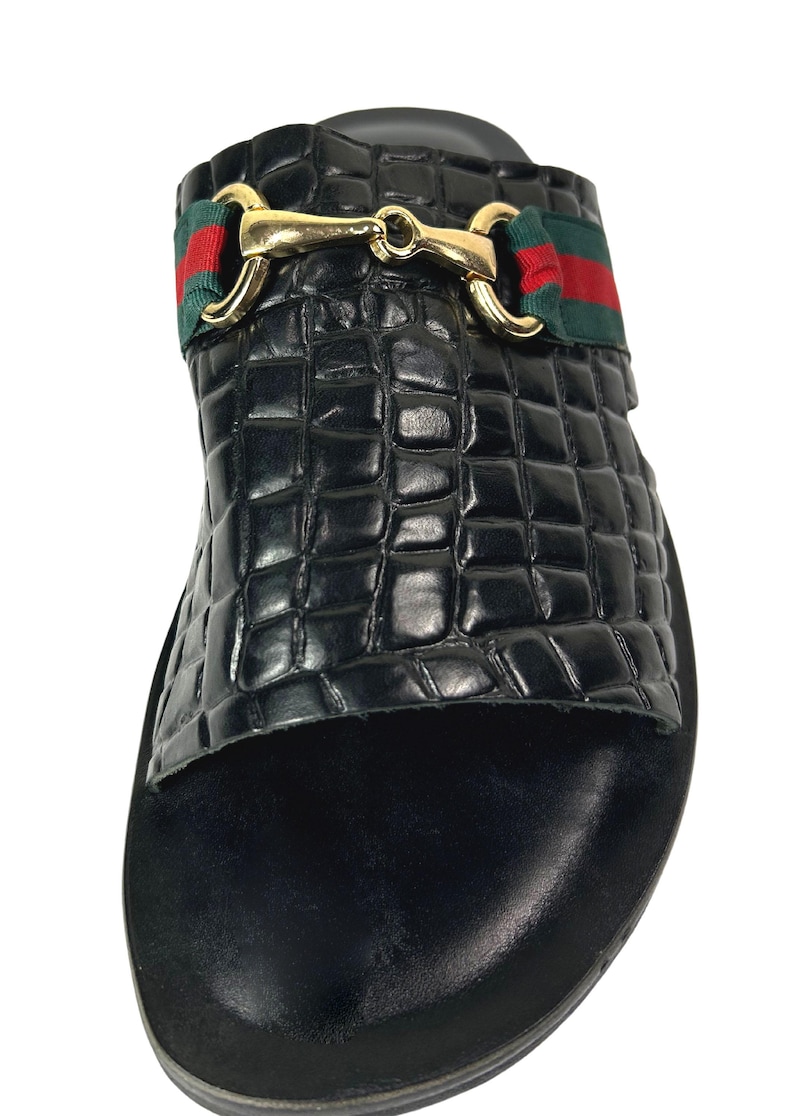 May include: A black slide sandal featuring a textured crocodile-embossed finish on the upper strap. The strap is accented with a horizontal green and red fabric band and a polished gold-tone metal horsebit hardware detail across the center.