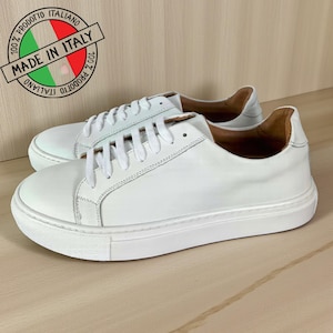 Könnte beinhalten: Ein Paar weiße Ledersneaker mit weißen Schnürsenkeln und Sohlen. Die Low-Top-Sneaker werden auf einer Holzoberfläche präsentiert. Das Bild enthält einen "Made in Italy"-Stempel.