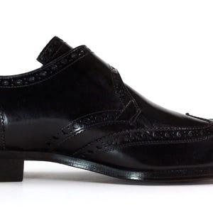 May include: A black leather oxford shoe featuring a wingtip design with decorative brogue perforations along the seams, a rounded toe, a low stacked heel, and a smooth polished finish on the exterior surface of the footwear.