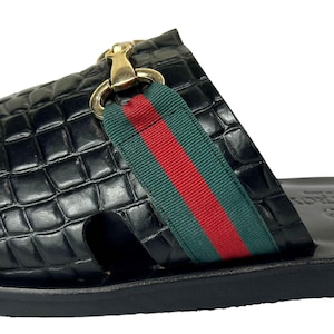 May include: A black slide sandal featuring a crocodile-embossed leather upper with a green and red striped fabric band across the top. The sandal includes a gold-toned metal horsebit detail and a smooth black leather footbed with a visible logo stamp.