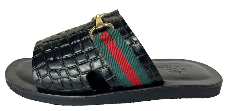 May include: A black slide sandal featuring a crocodile-embossed leather upper with a green and red striped fabric band across the top. The sandal includes a gold-toned metal horsebit detail and a smooth black leather footbed with a visible logo stamp.