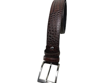 Può includere: Una cintura in pelle marrone con un motivo strutturato a forma di coccodrillo. La cintura ha una fibbia rettangolare color argento e un passante abbinato. La pelle sembra essere di una tonalità marrone scuro.